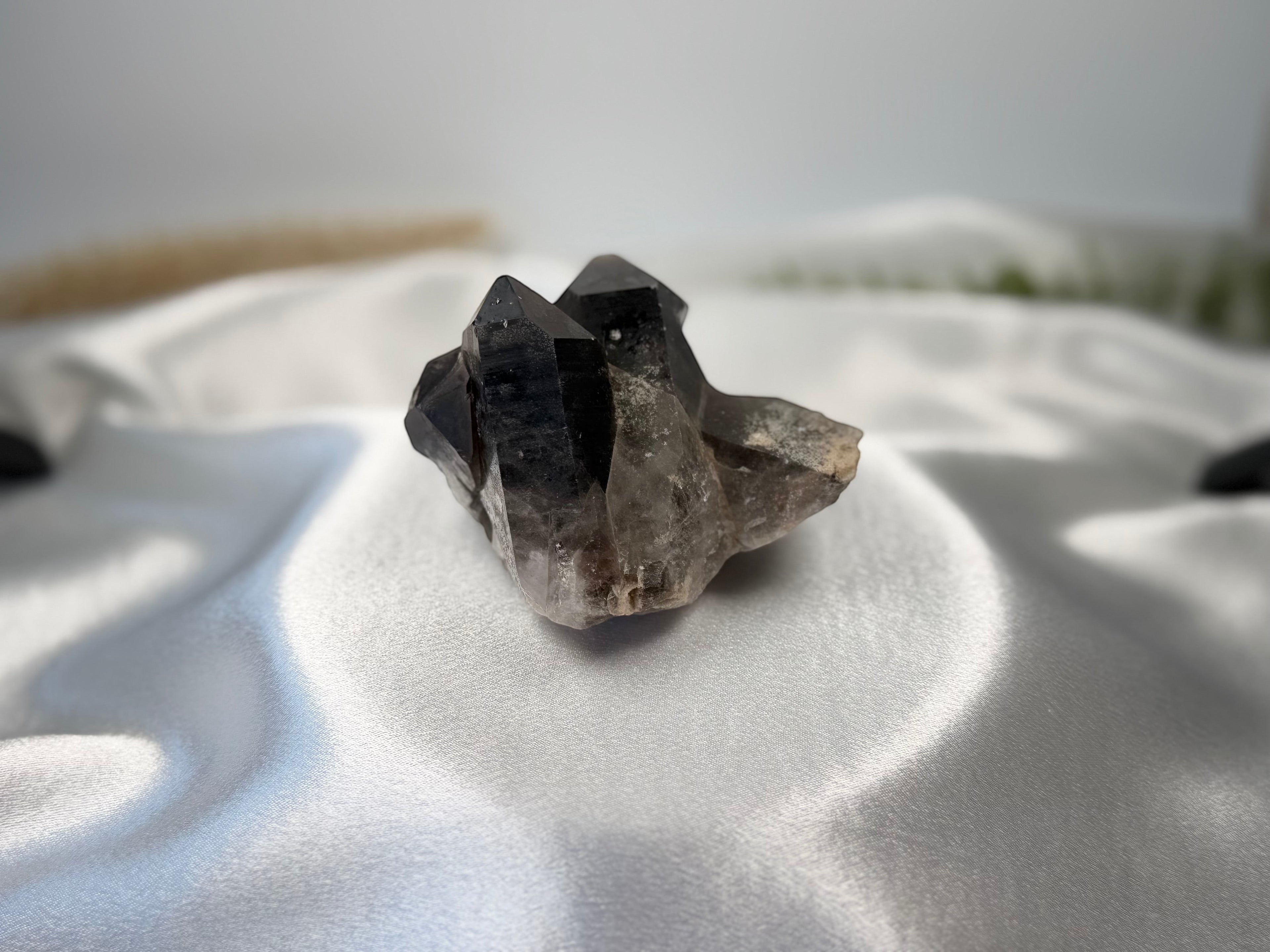 Treasured Energies raw smoky quartz crystal cluster with deep brown and grey tones on a white satin background.