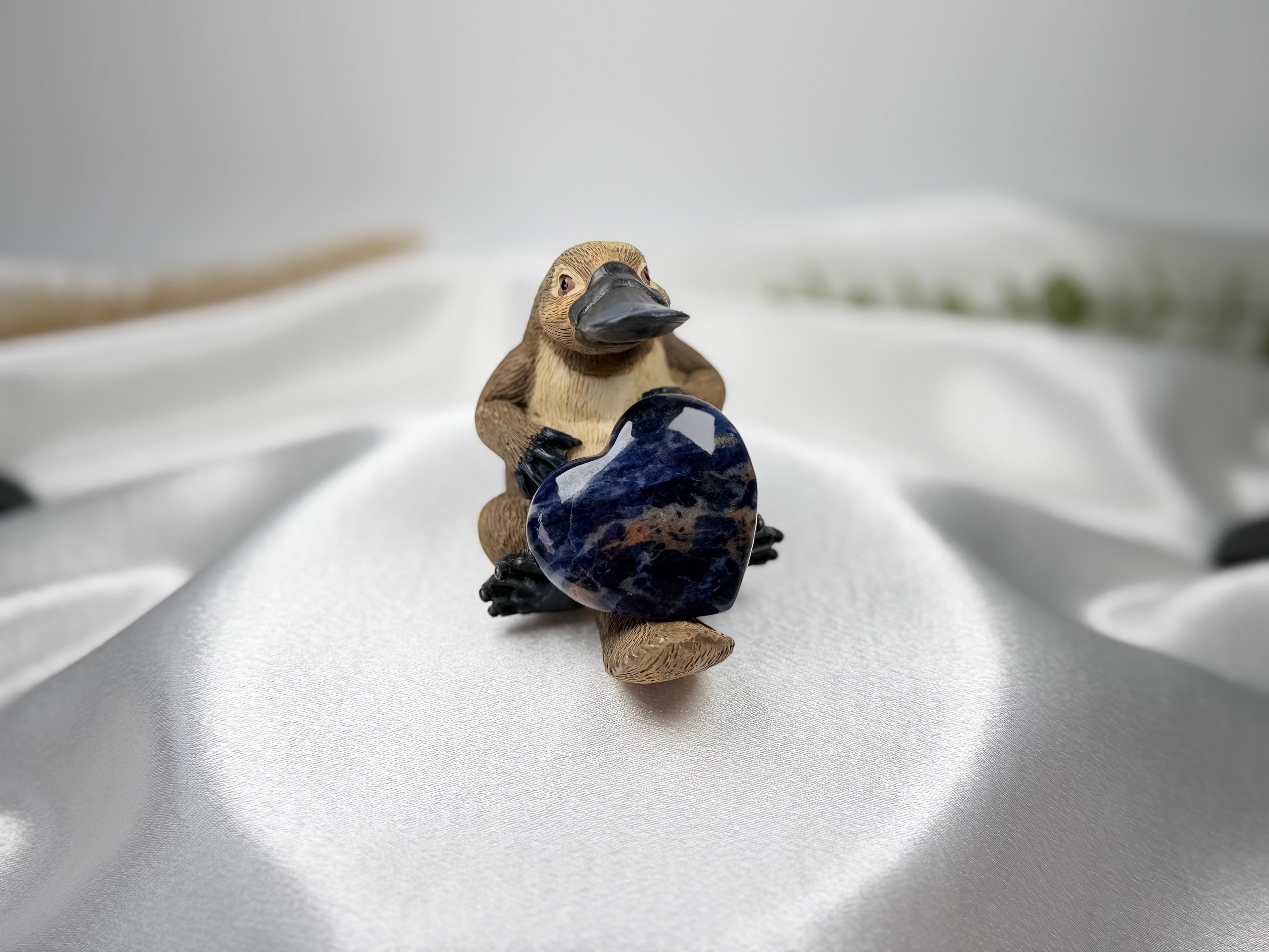Treasured Energies polished blue sodalite crystal heart held by a small platypus figurine on white satin fabric.