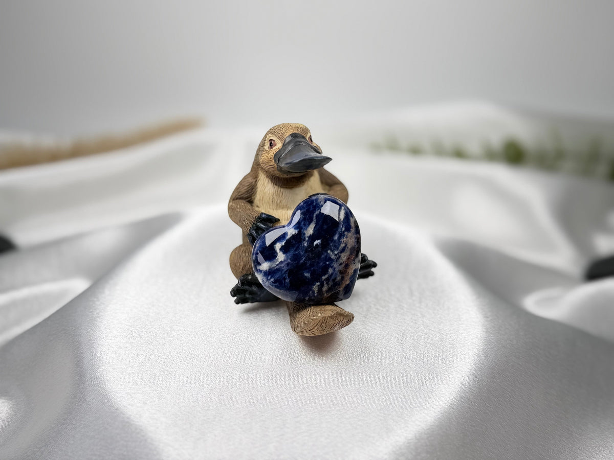Treasured Energies polished blue sodalite crystal heart held by a small platypus figurine on white satin fabric.