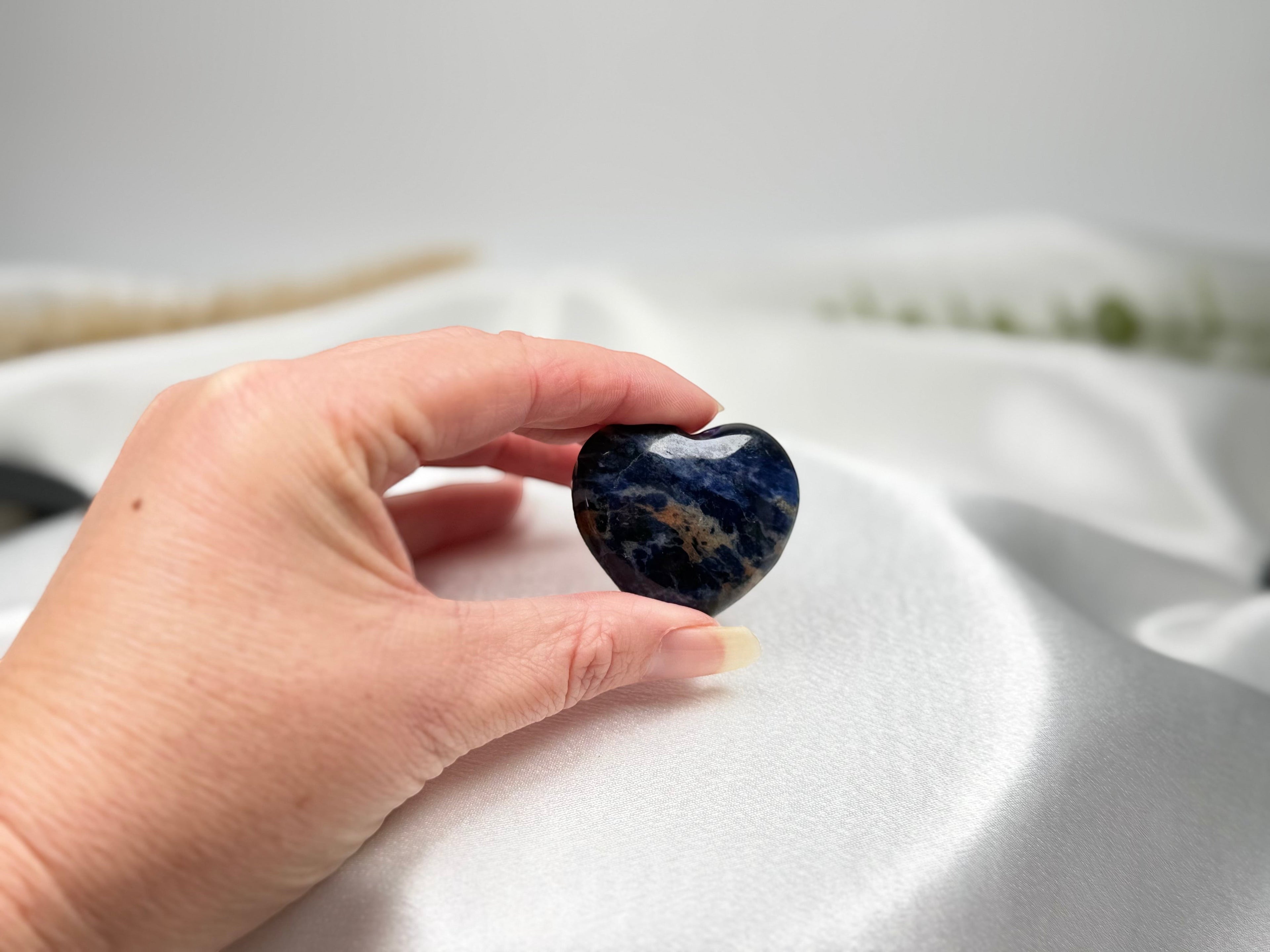 Treasured Energies polished blue sodalite crystal heart held in a hand against a soft white satin background.