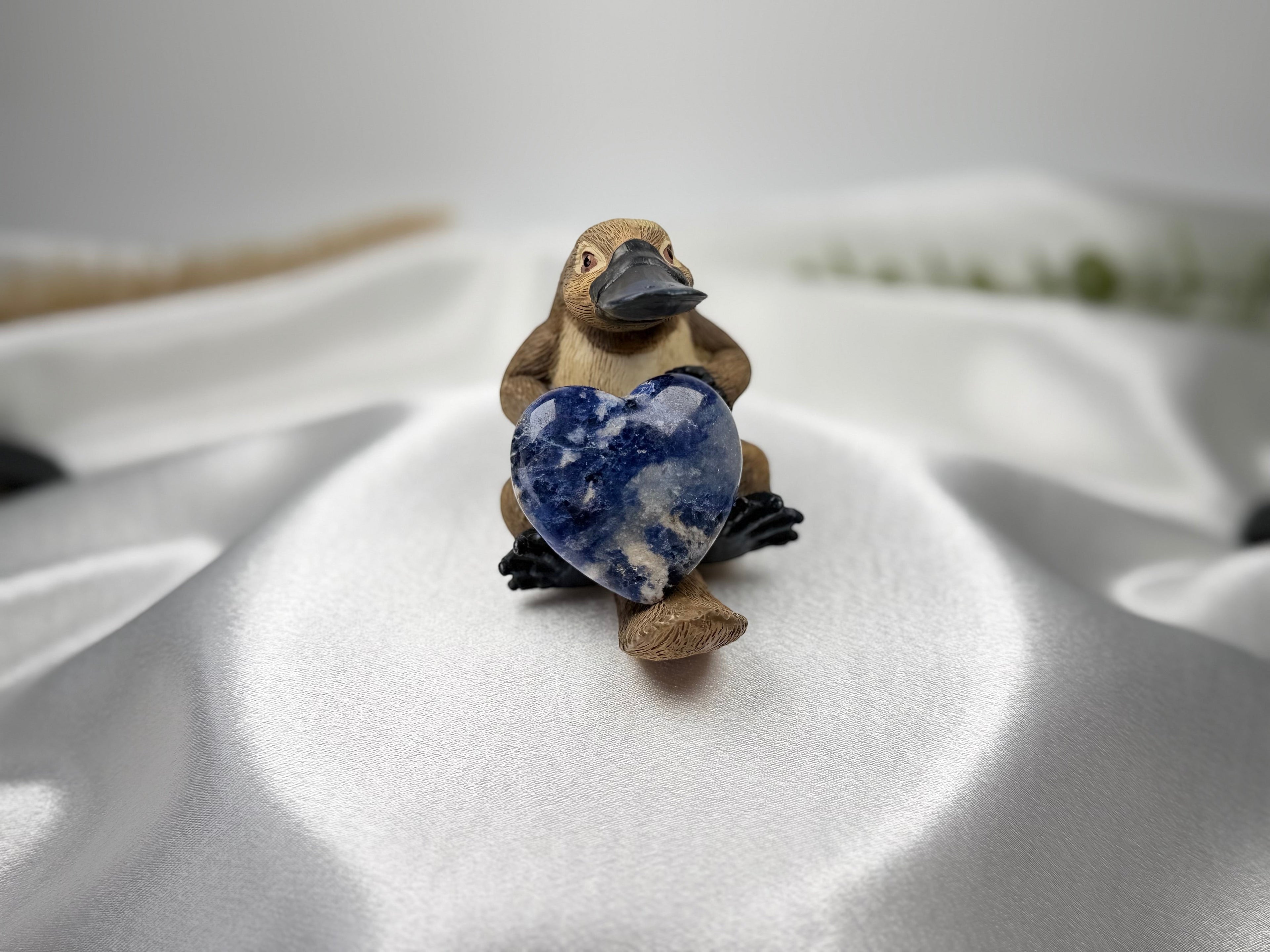 Treasured Energies polished blue and white sodalite crystal heart held by a small platypus figurine on white fabric.