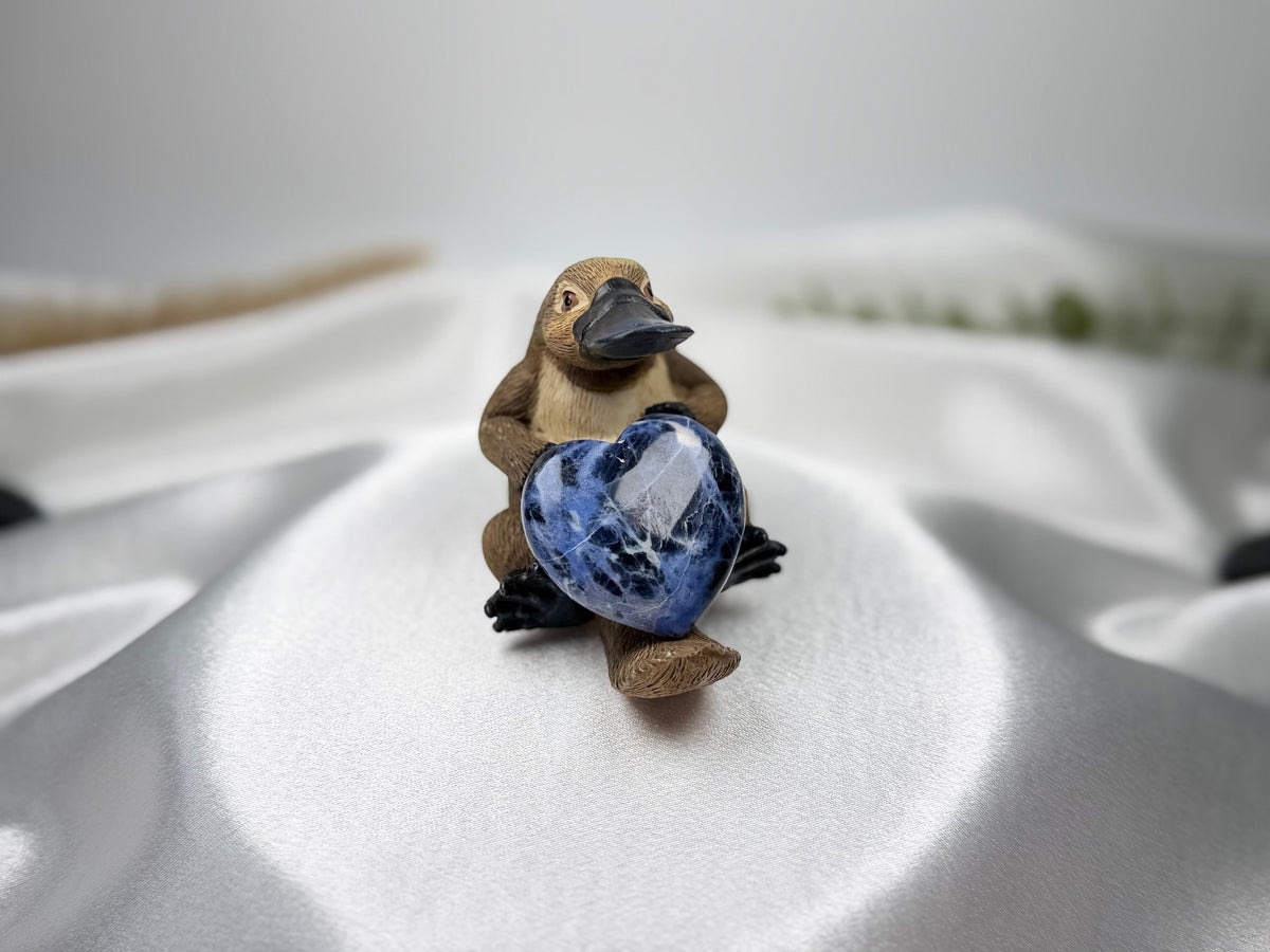 Treasured Energies polished blue sodalite crystal heart held by a small platypus figurine on white satin fabric