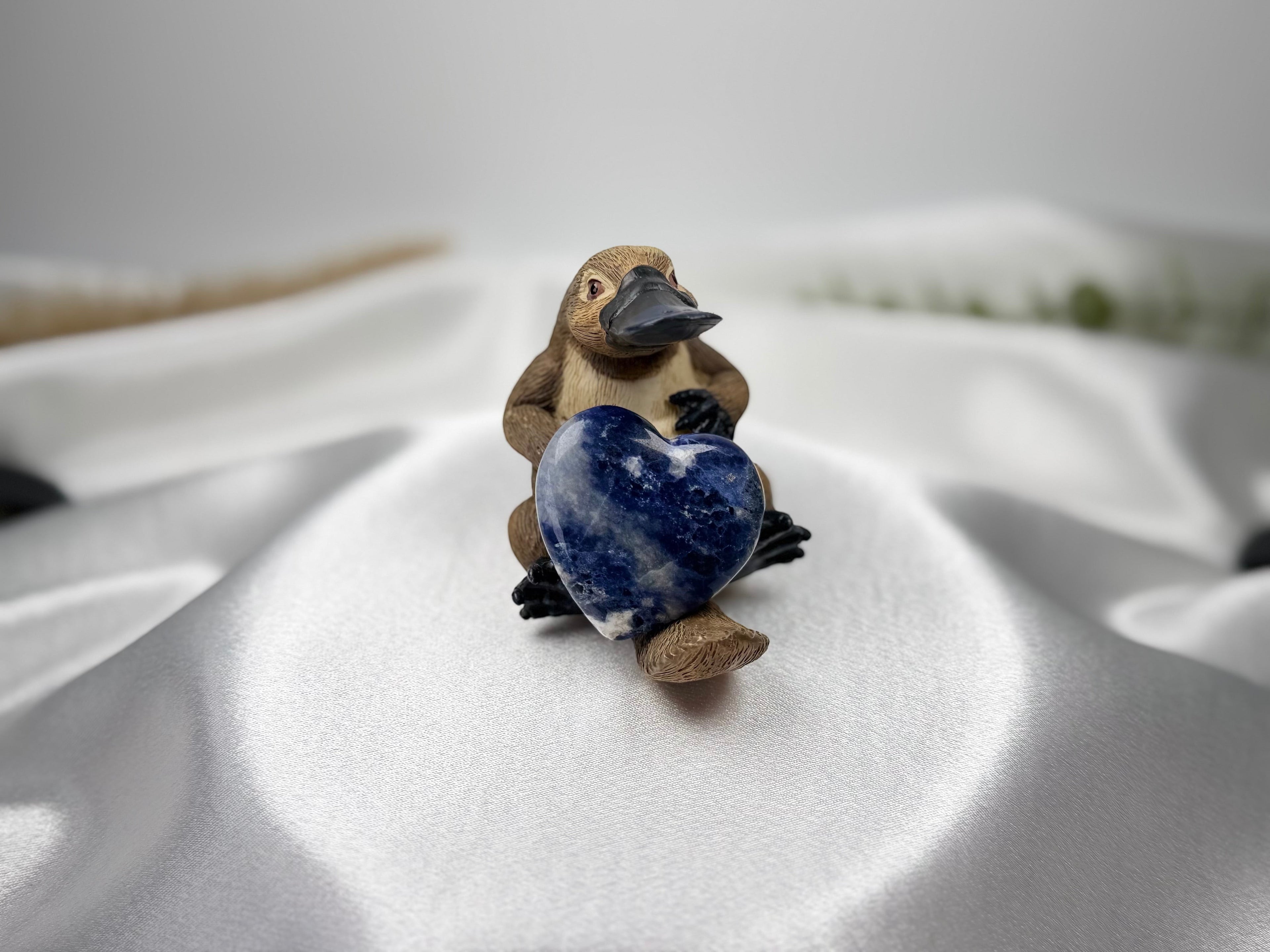 Treasured Energies polished blue Sodalite crystal heart held by a small platypus figurine on white satin fabric