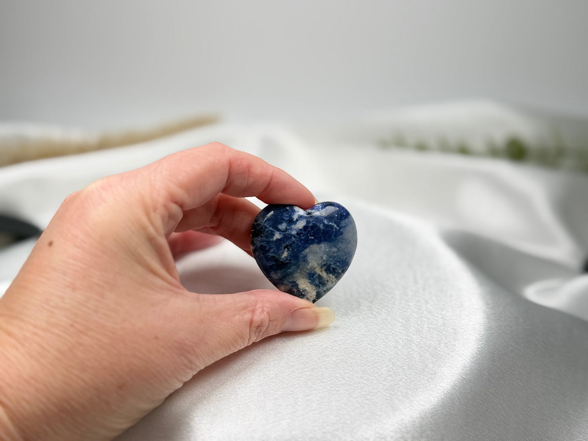 Treasured Energies polished blue and white Sodalite crystal heart held in a hand against a soft white satin background.