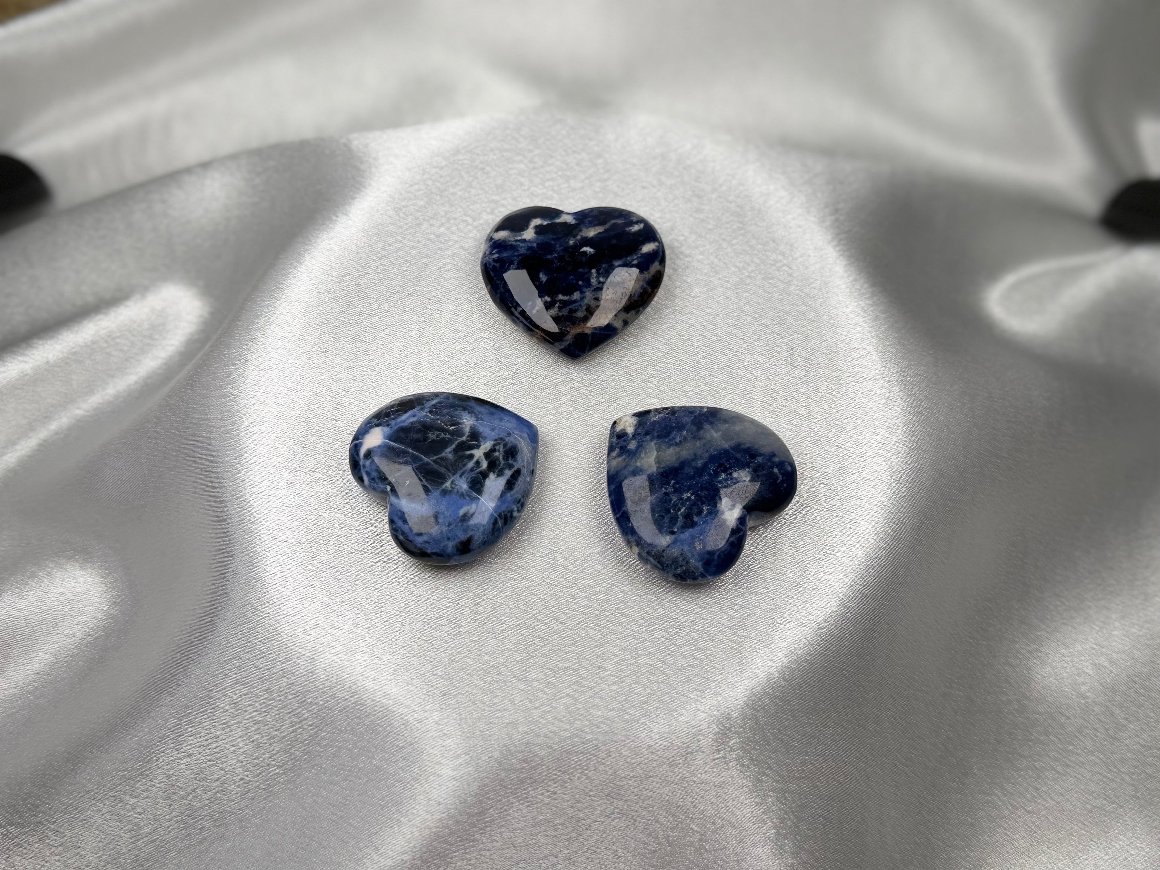Three polished Treasured Energies sodalite crystal hearts with deep blue and white patterns on a satin fabric.