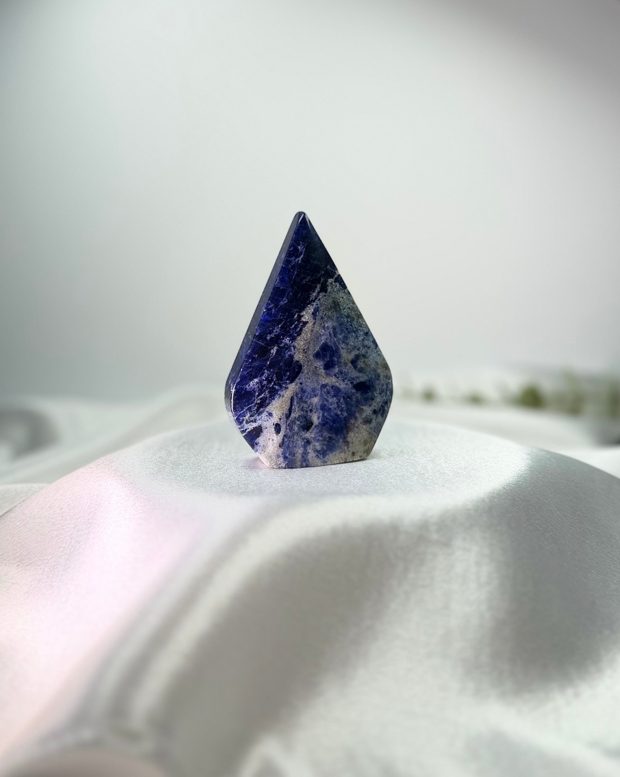 Treasured Energies polished blue sodalite crystal flame freeform with white mineral inclusions on a white satin base.