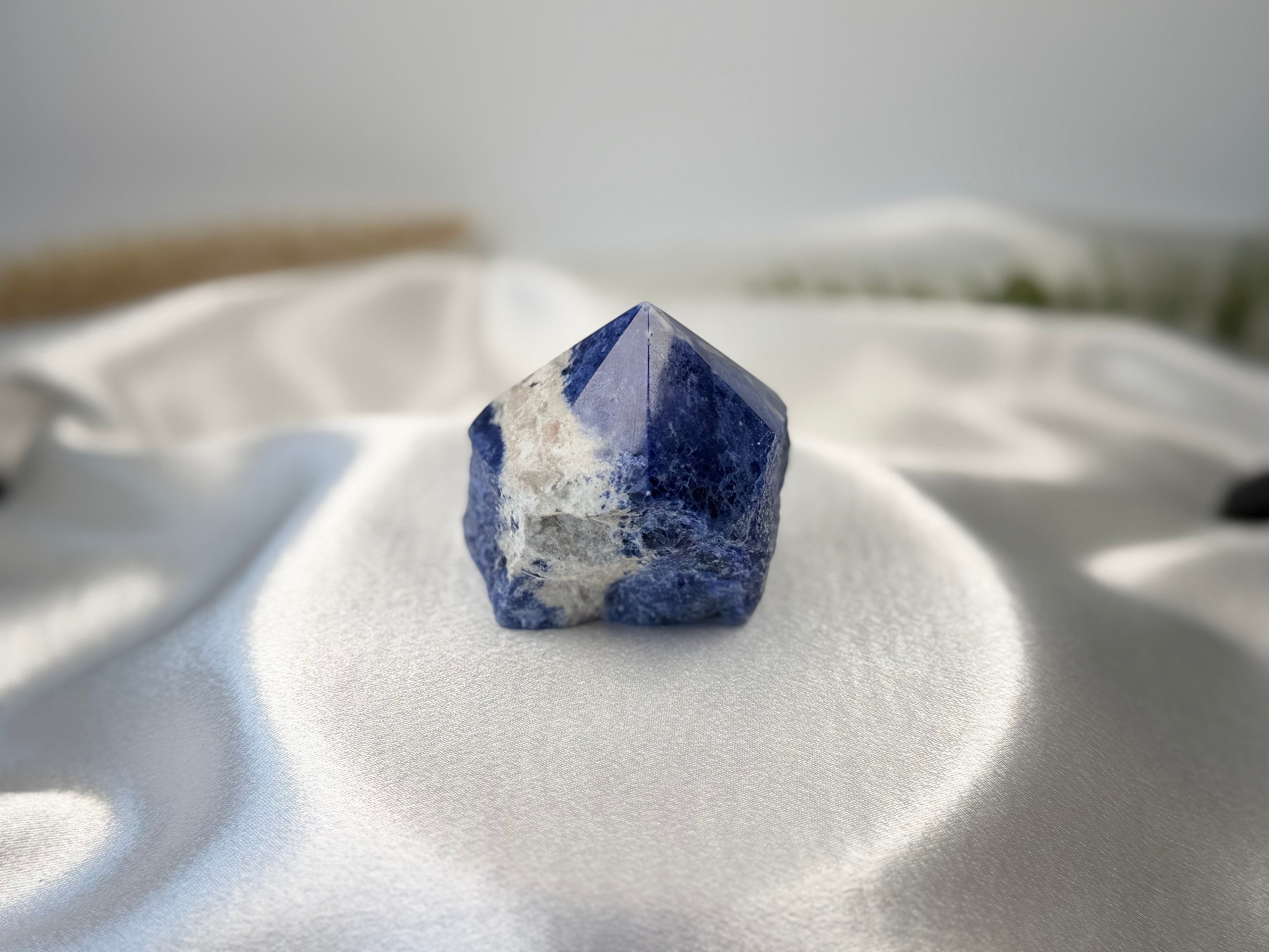Treasured Energies sodalite crystal tower with a polished point and raw base on a white satin fabric background.