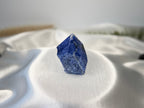 Treasured Energies royal blue Sodalite crystal point with a polished tip and raw base on a white satin fabric background.