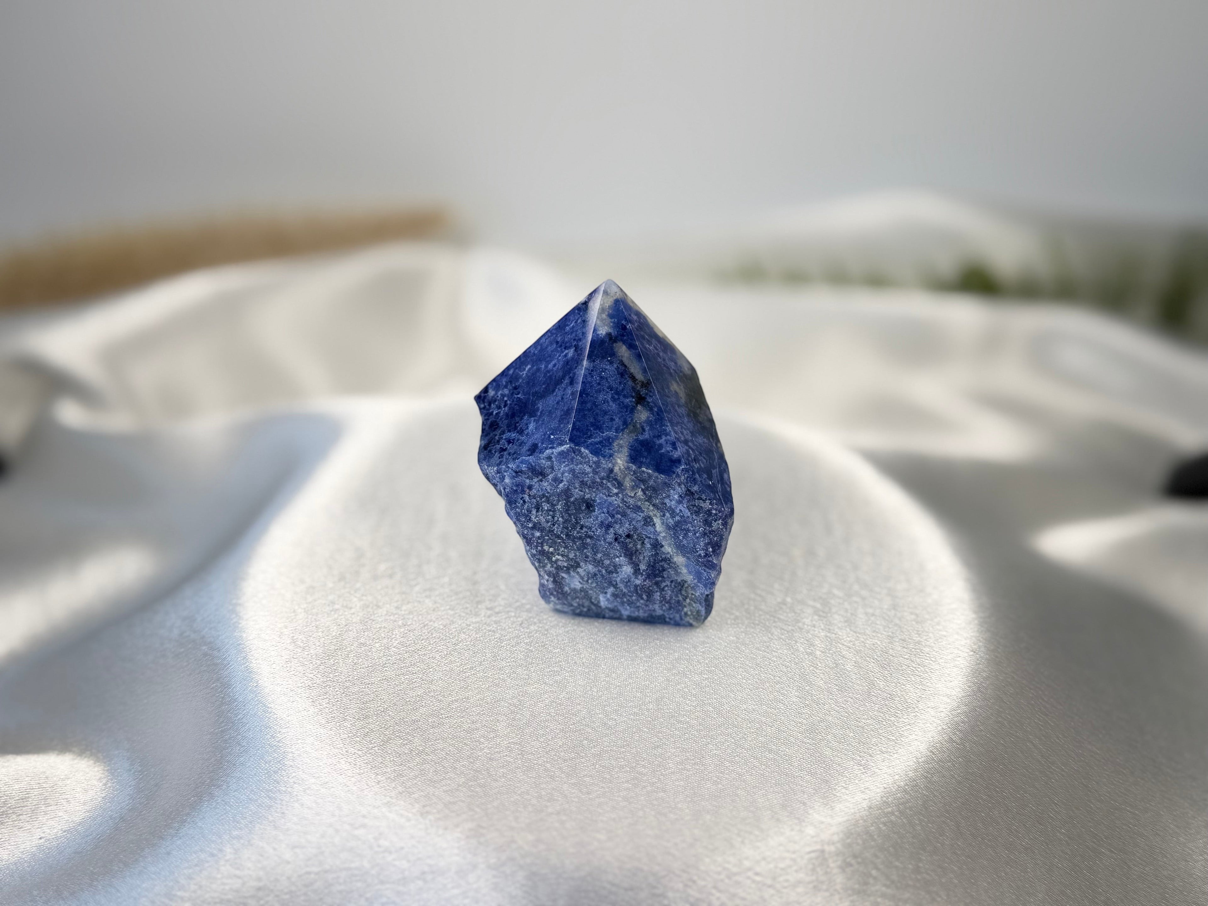 Treasured Energies royal blue Sodalite crystal point with a polished tip and raw base on a white satin fabric background.