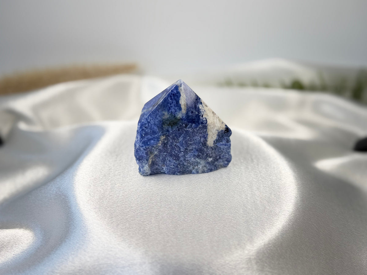 Treasured Energies Sodalite crystal point with a polished top and raw, natural base on a white satin background.