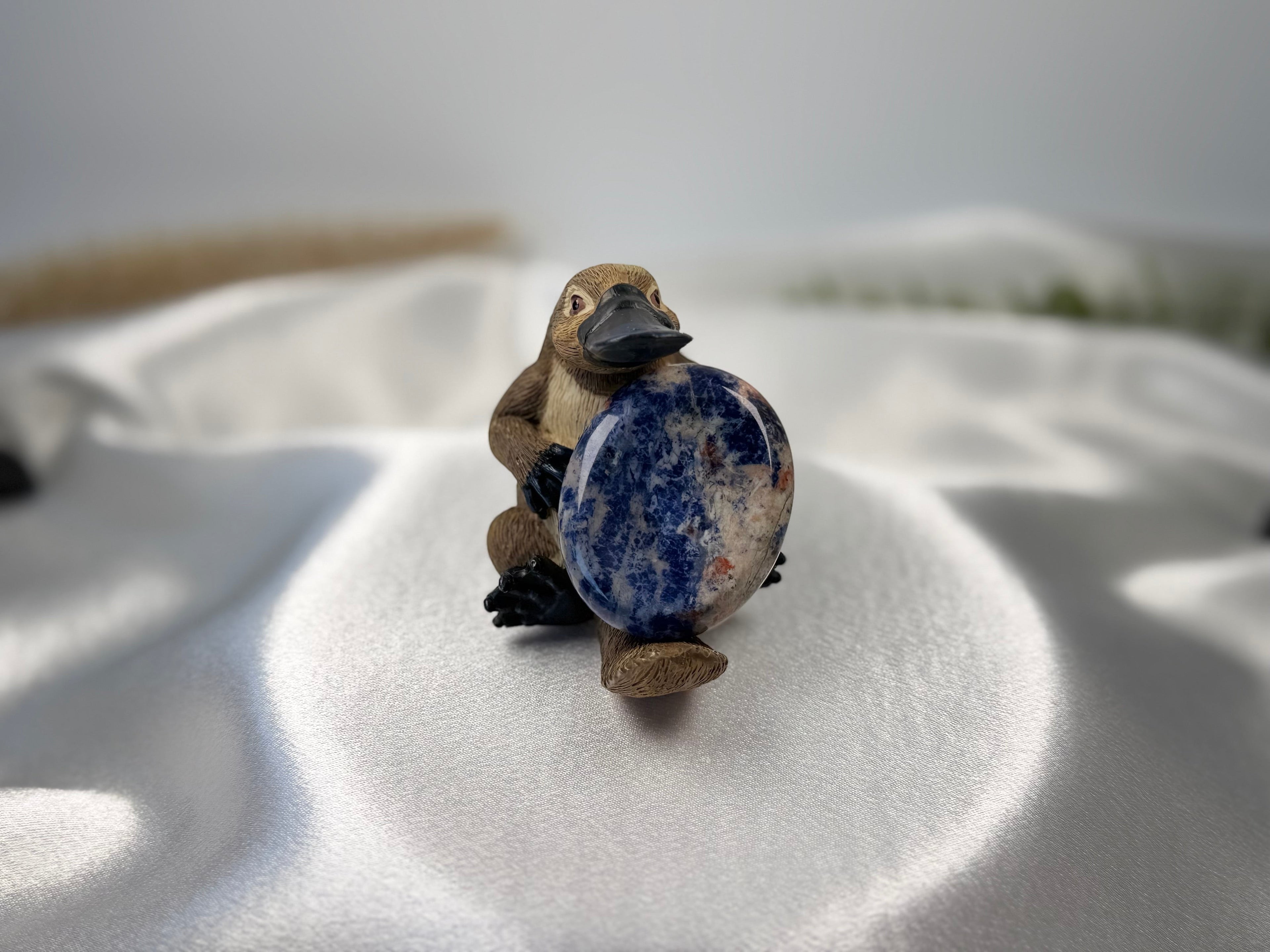 Treasured Energies blue and white Sodalite worry stone held by a small platypus figurine on a white satin fabric