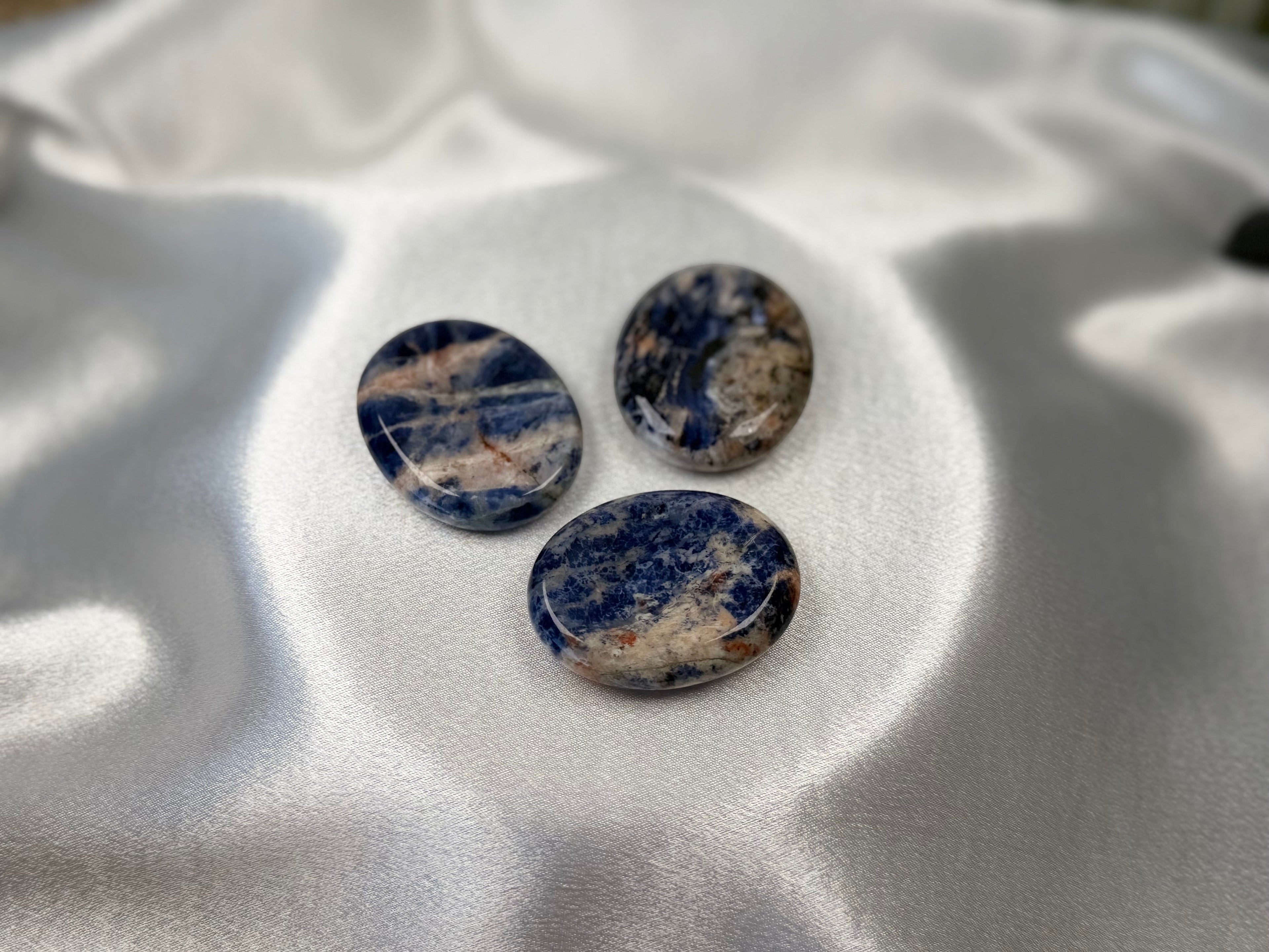 Treasured Energies smooth oval sodalite worry stones with deep blue and white mineral patterns on white satin fabric.