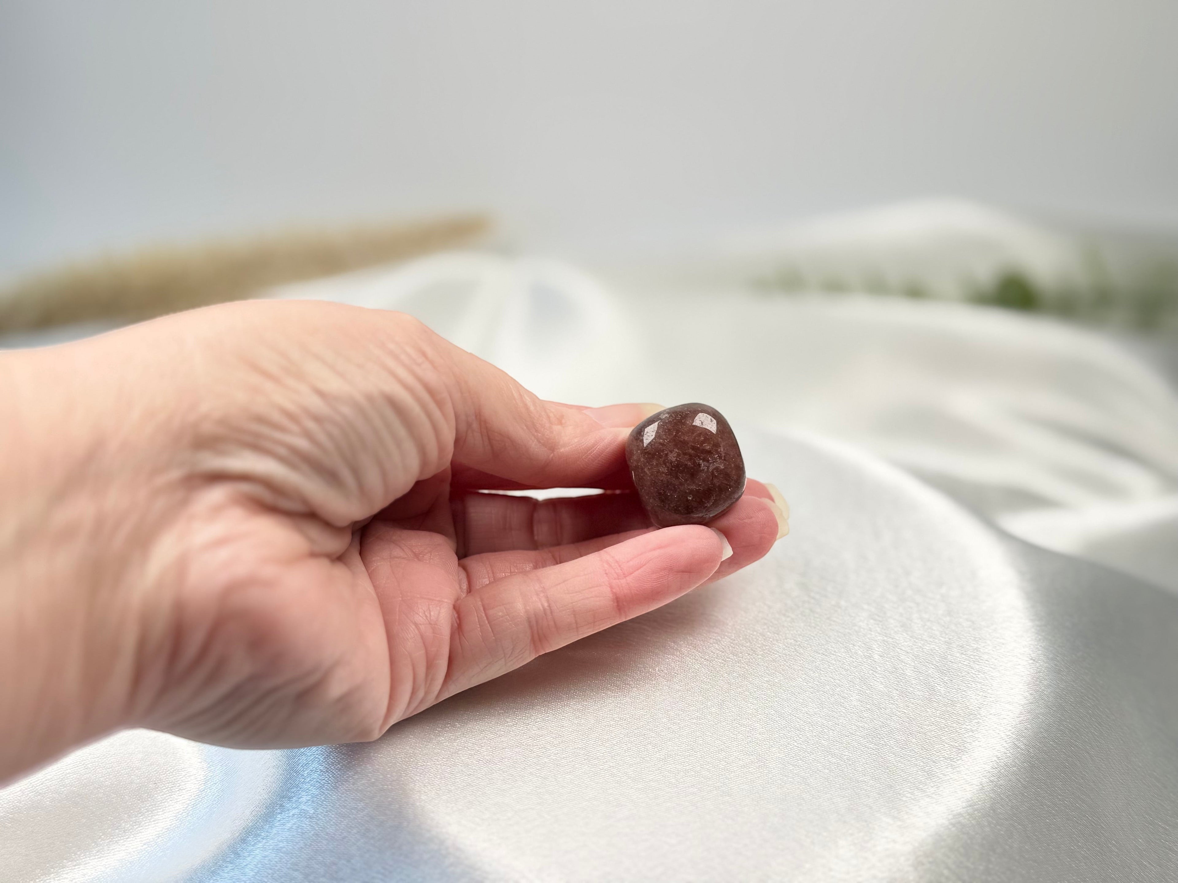 Treasured Energies polished strawberry quartz tumbled crystal held in a hand against a soft white satin background