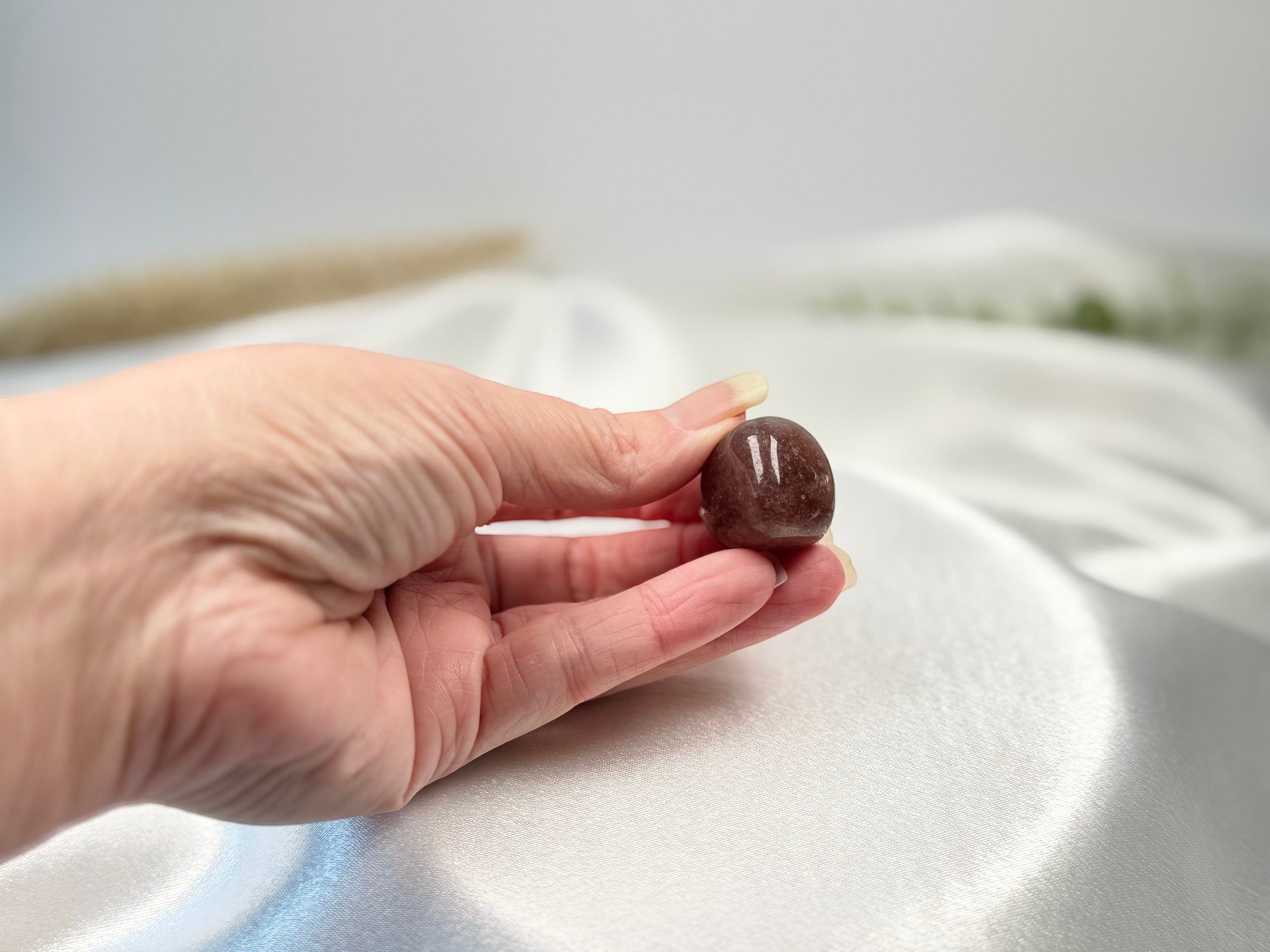 Treasured Energies polished Strawberry Quartz tumbled crystal held in a hand against a soft white satin background.