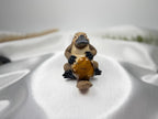 Treasured Energies polished tiger eye crystal heart held by a small brown platypus figurine on white satin fabric