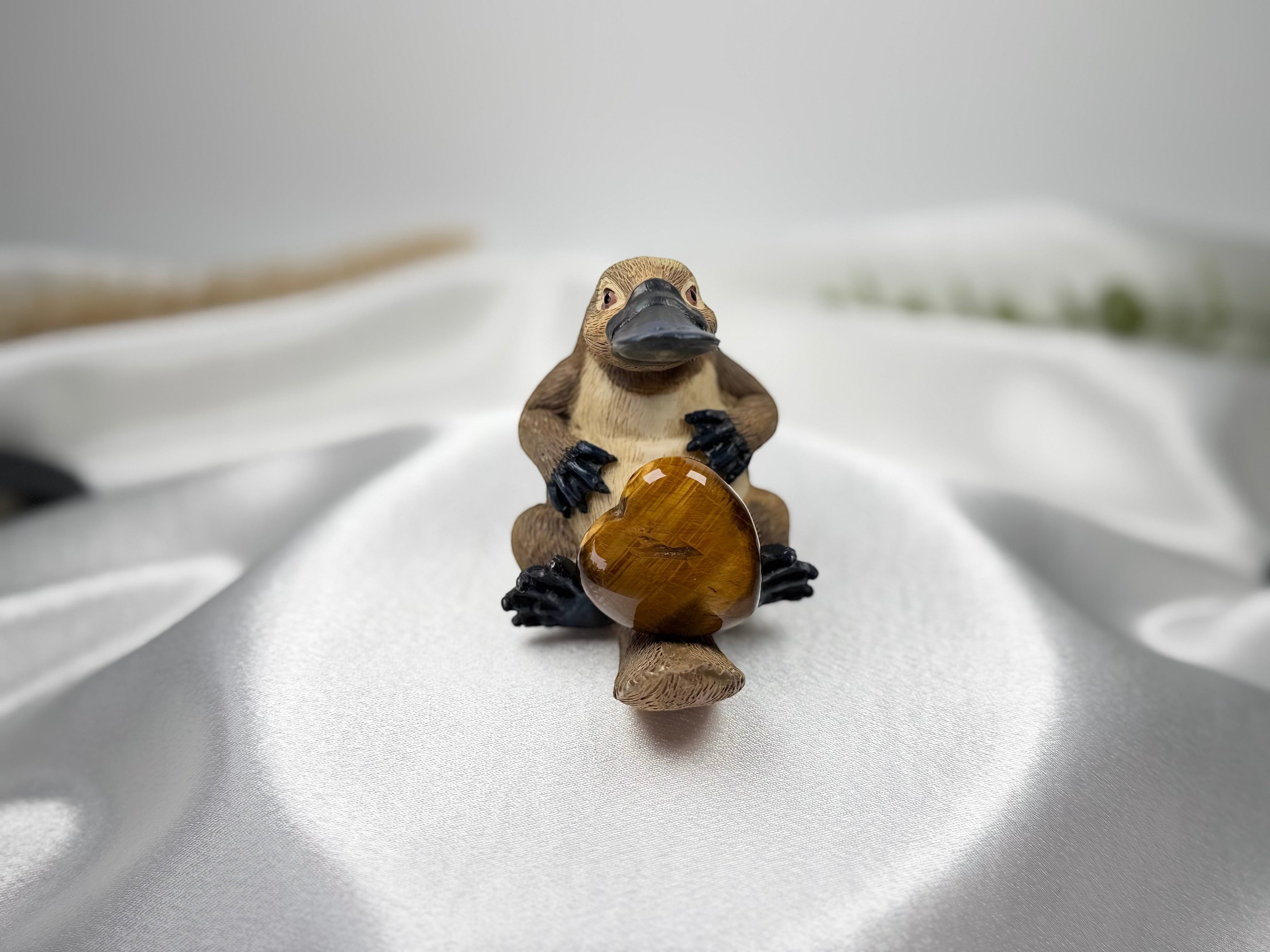 Treasured Energies polished tiger eye crystal heart held by a small brown platypus figurine on white satin fabric
