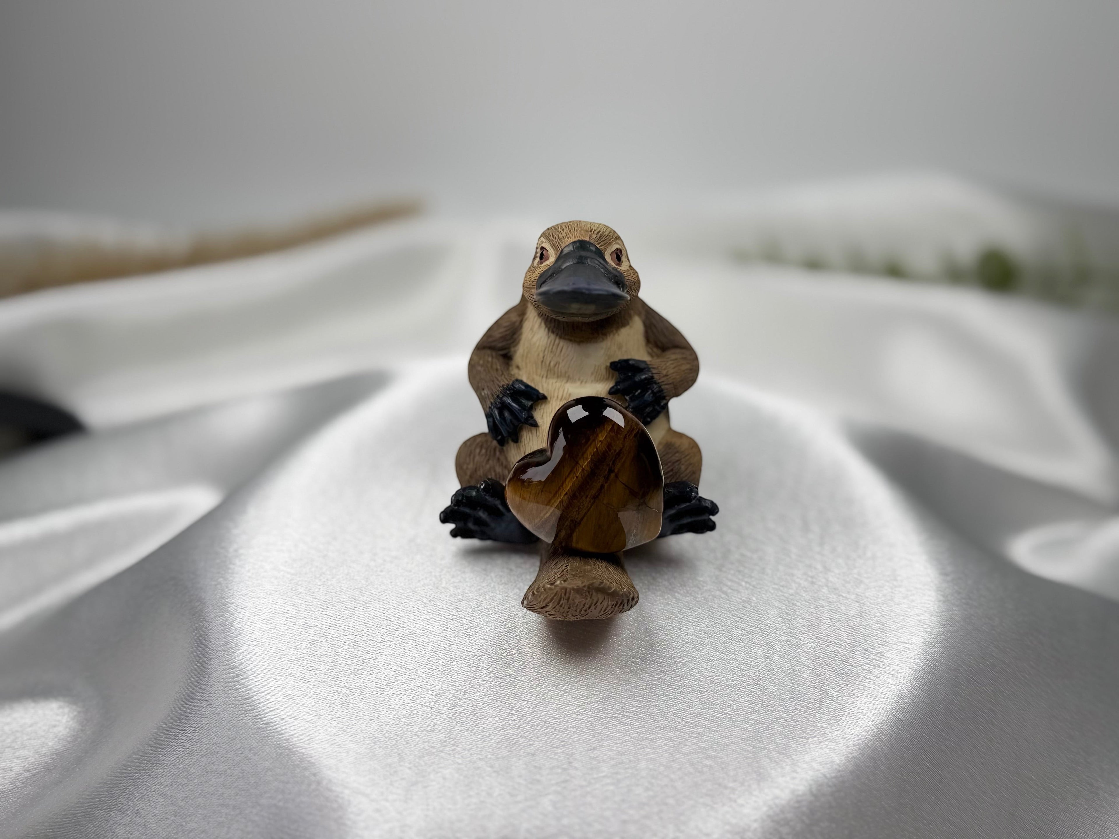 Treasured Energies polished brown tiger eye crystal heart held by a small carved platypus figurine on white fabric.