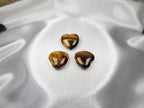 Three polished Treasured Energies Tiger Eye crystal hearts with golden-brown chatoyant shimmer on white satin fabric.
