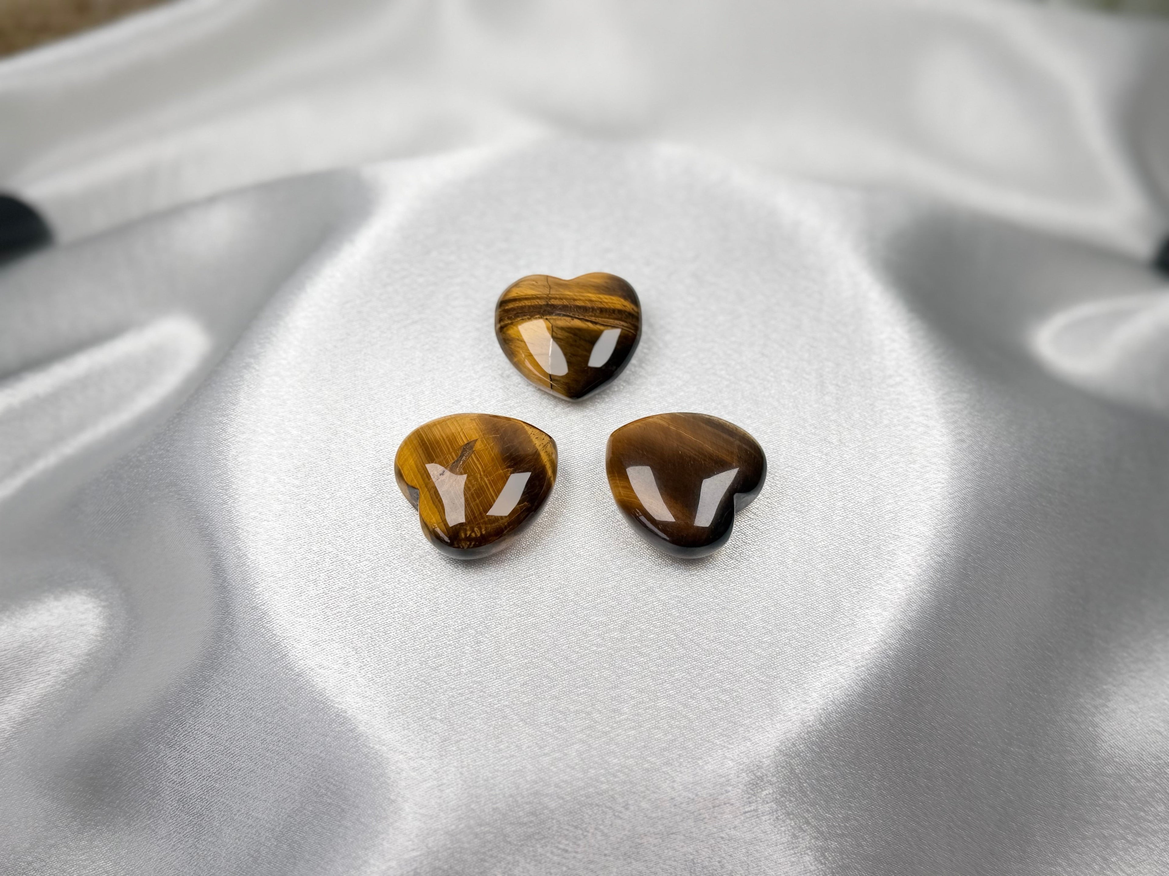 Three polished Treasured Energies Tiger Eye crystal hearts with golden-brown chatoyant shimmer on white satin fabric.