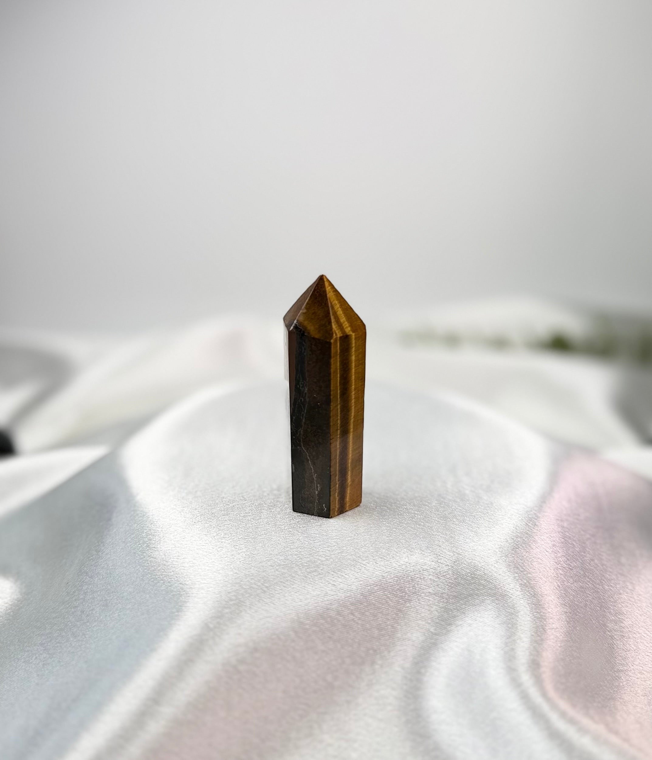 Treasured Energies Tigers Eye crystal tower with golden-brown chatoyant bands on a soft white satin fabric background.