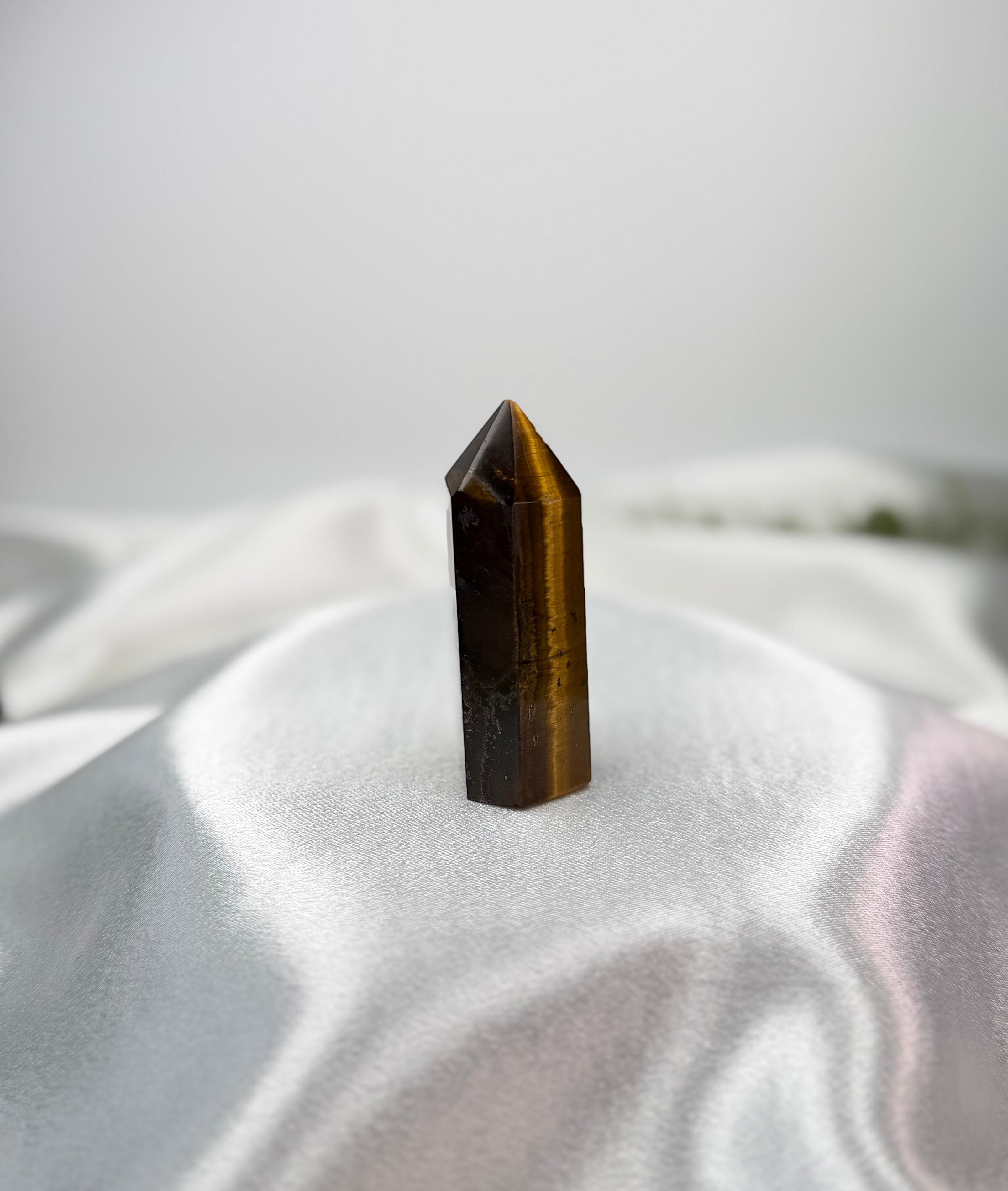 Treasured Energies Tigers Eye crystal tower with golden-brown chatoyant bands on a white satin fabric background.