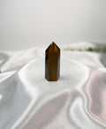 Treasured Energies polished Tigers Eye crystal tower with a pointed tip on white satin fabric.