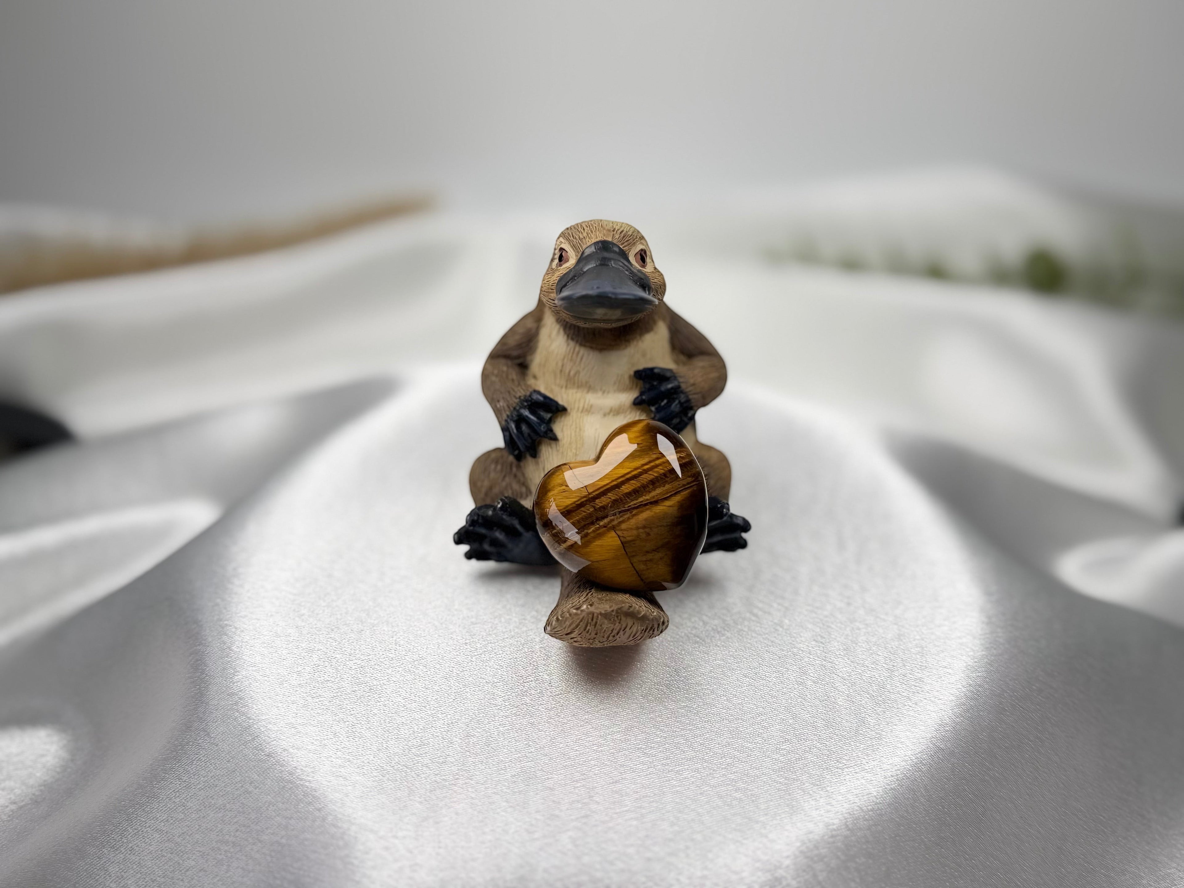 Treasured Energies polished Tigers Eye crystal heart held by a small platypus figurine on a white satin background.