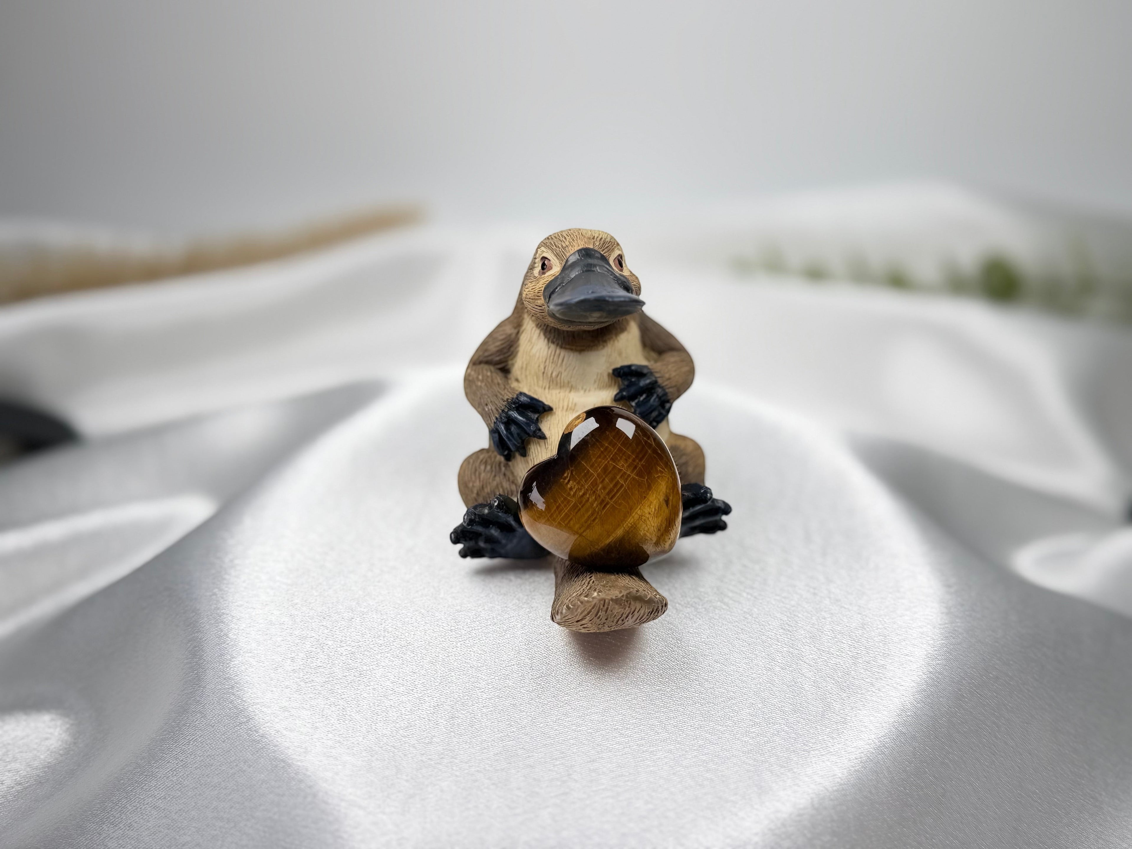 Treasured Energies polished Tigers Eye crystal heart held by a small brown platypus figurine on white satin fabric.