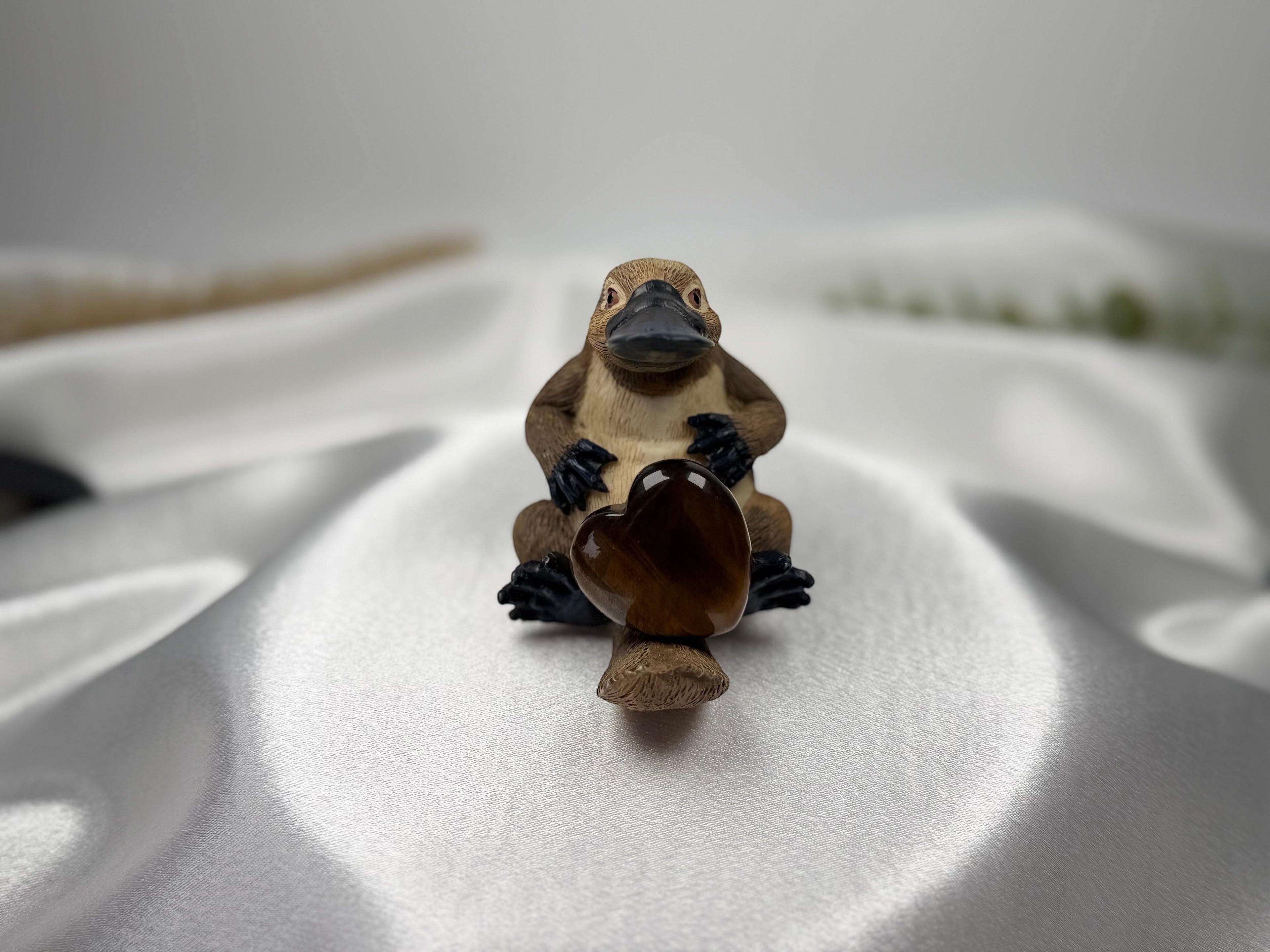 Treasured Energies heart-shaped Tiger Eye crystal held by a small, detailed platypus figurine on white satin fabric.