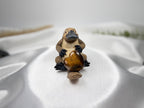 Treasured Energies heart-shaped polished Tiger Eye crystal held by a small brown platypus figurine on white silk.