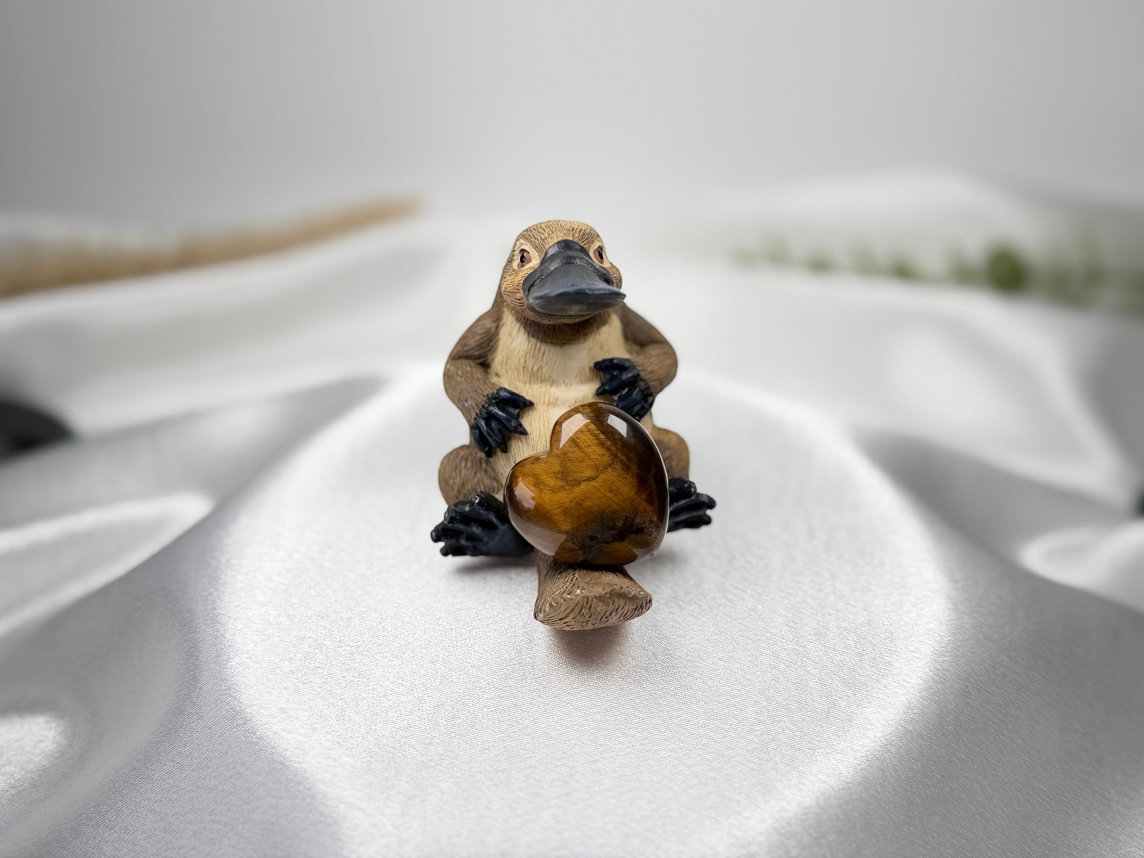 Treasured Energies polished brown tiger eye crystal heart held by a small platypus figurine on white satin fabric.