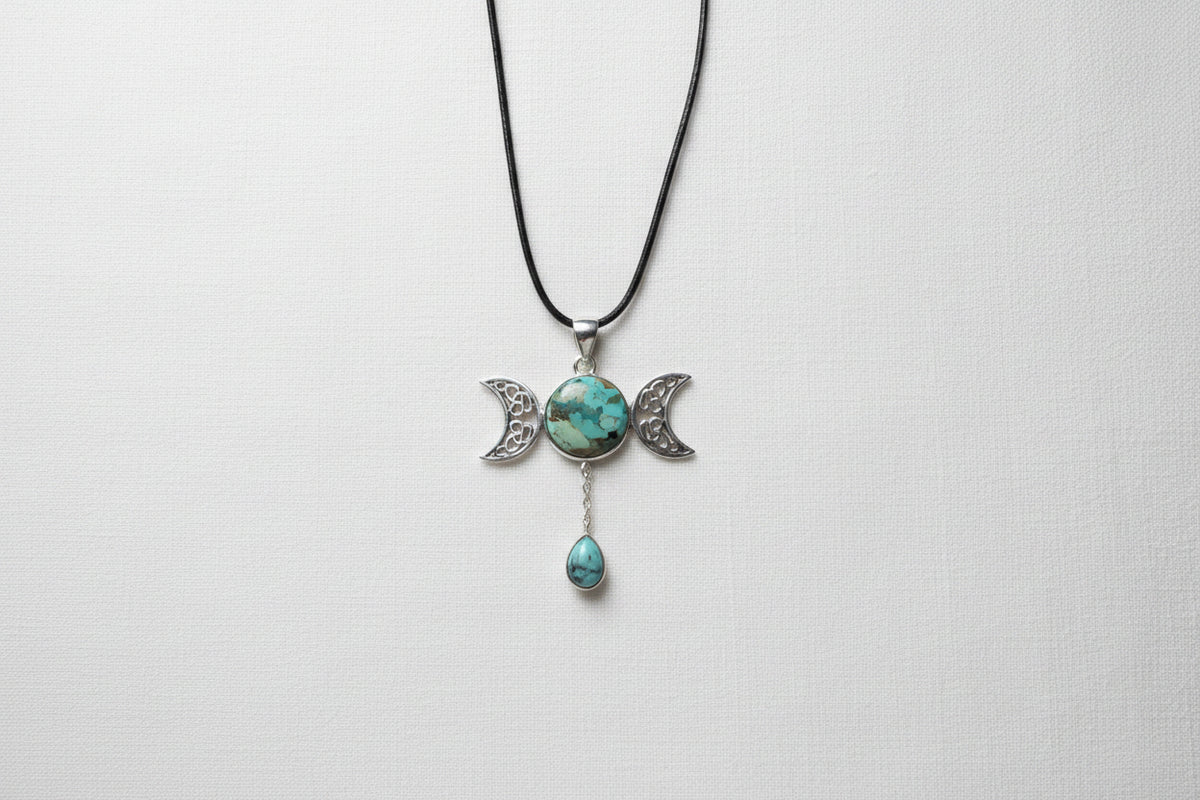 Treasured Energies Triple Moon pendant necklace with turquoise stones on a black cord against a textured grey fabric.