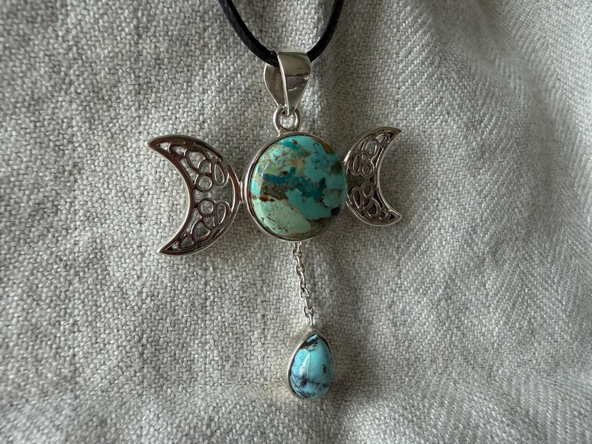 Treasured Energies Triple Moon Goddess pendant necklace with turquoise stones on a black cord against textured fabric