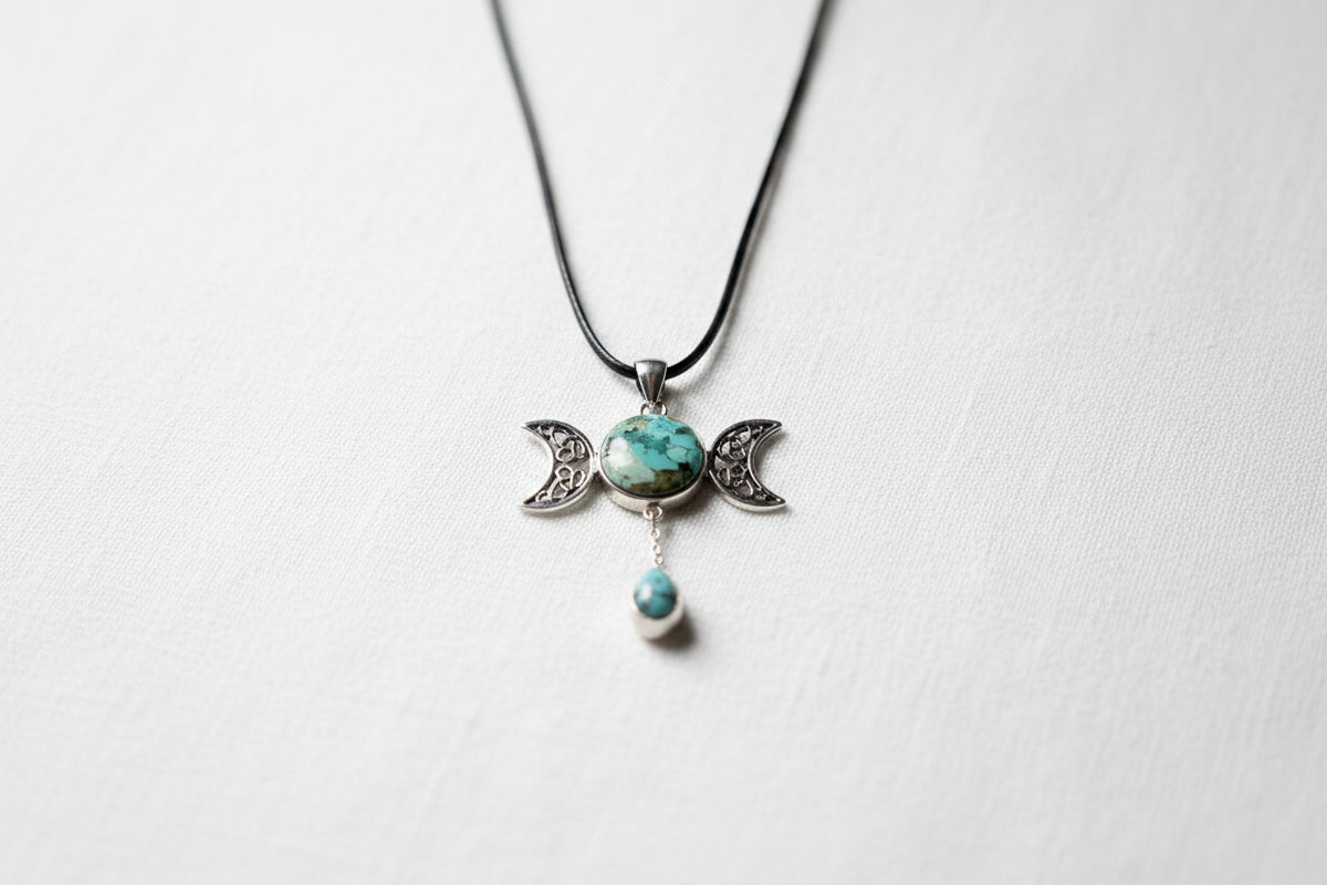 Treasured Energies Triple Moon pendant necklace with turquoise stones on a black cord against a textured grey fabric.