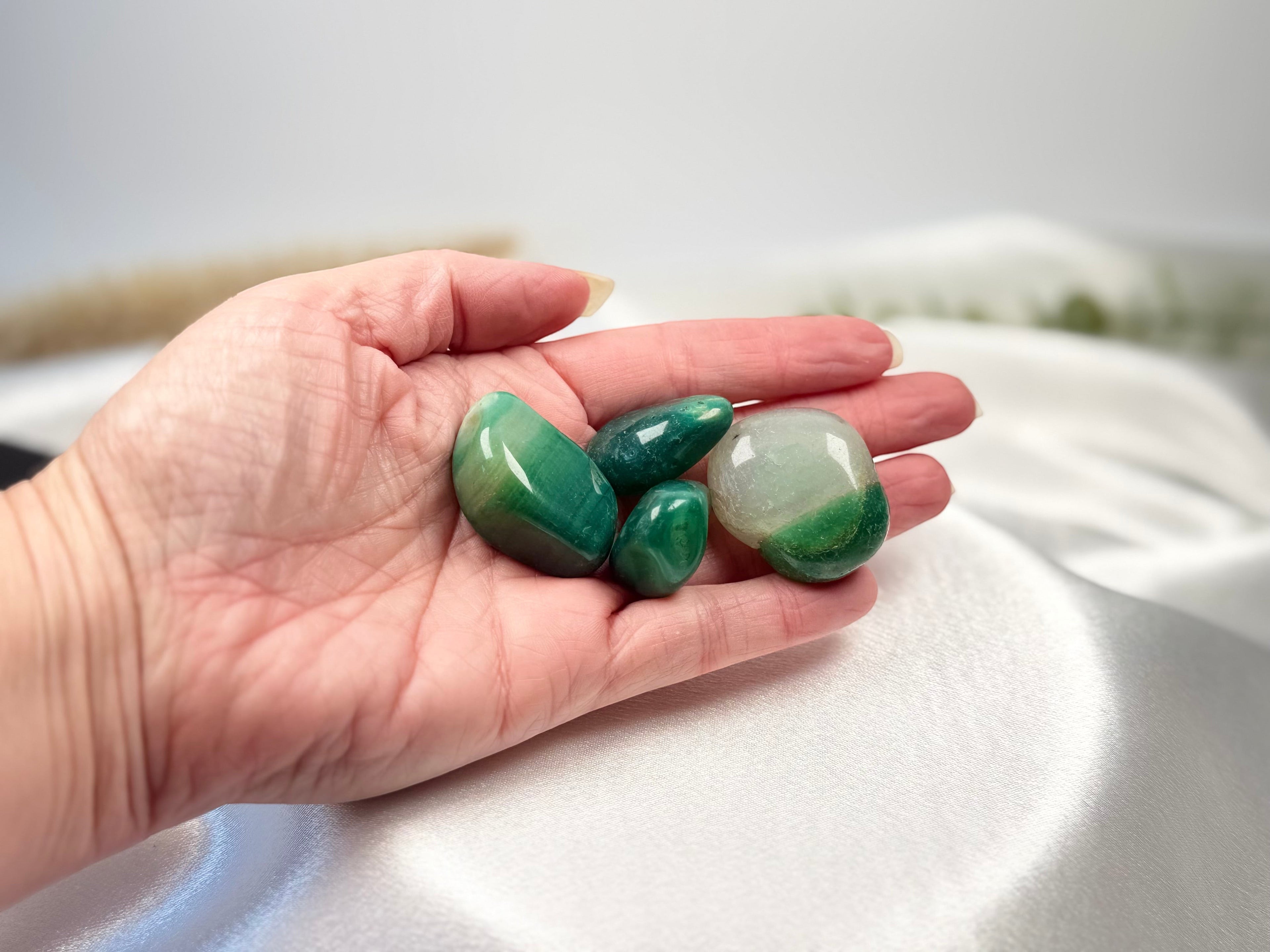 Treasured Energies tumbled Amazonite crystals in various shades of green held in a palm against a white silk fabric.