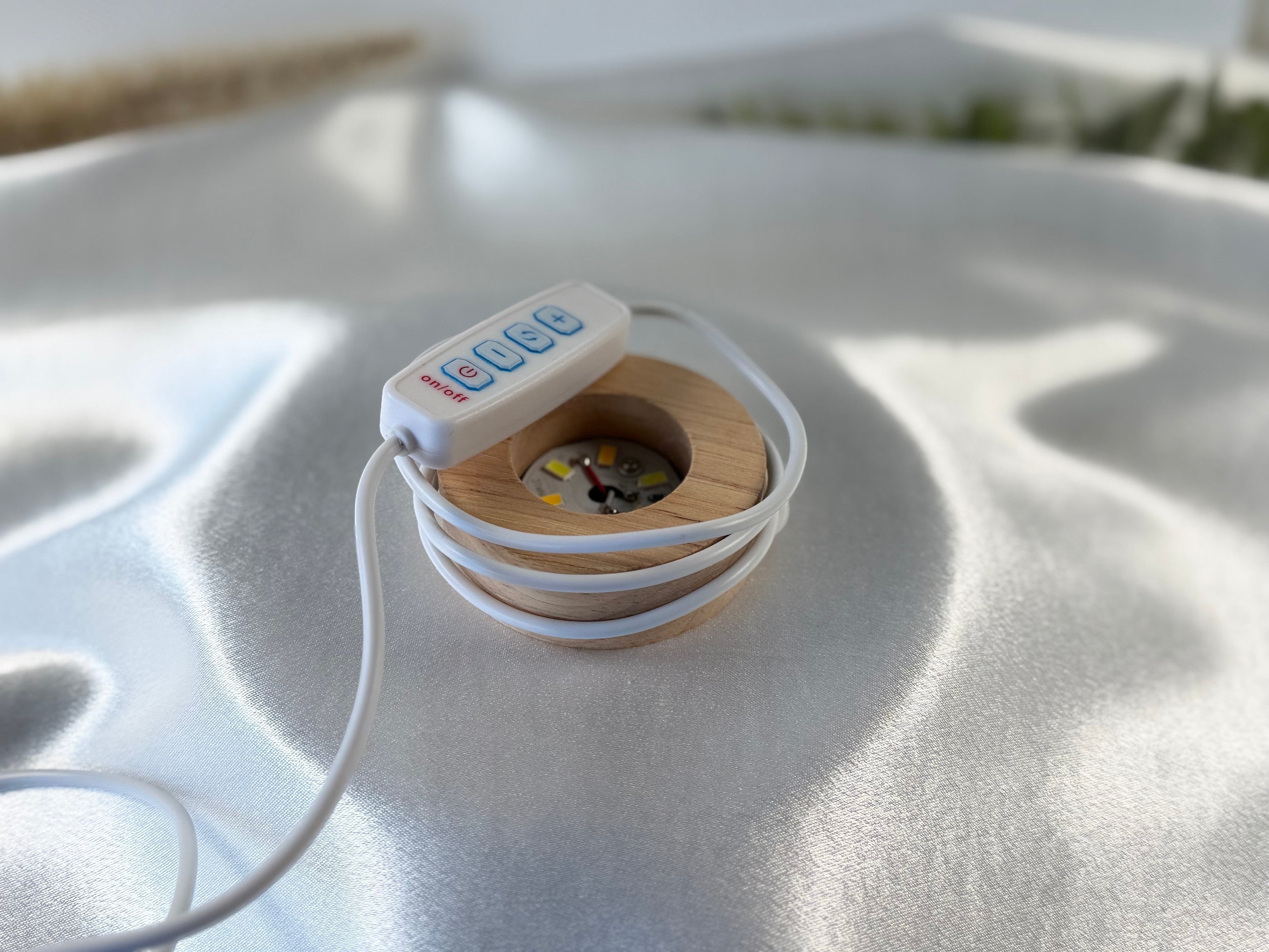 Treasured Energies small round wooden LED lamp base with white cord and control switch on a satin fabric background